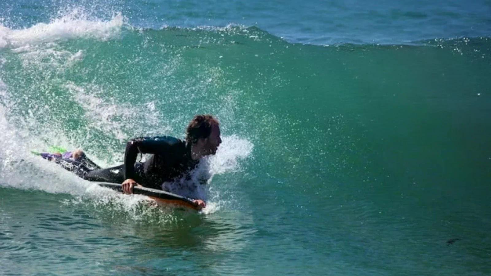 Surfer liegt in einer Welle auf einem Surfbrett, der Oberkörper aus dem Wasser gestützt, während die grüne Welle vor ihm breitet.
