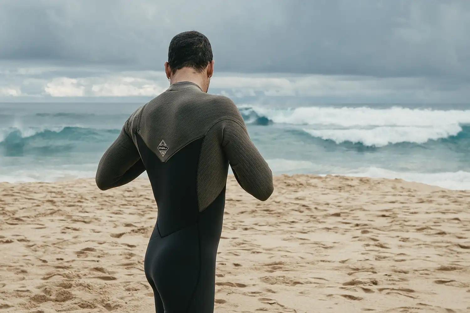 Surfer packt einen Neoprenanzug am Strand ein, Blick aufs Meer mit Wellen; grauer Himmel.