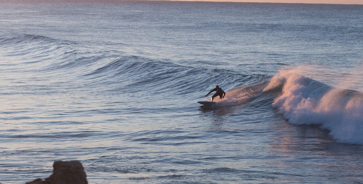 Surfer reitet eine Welle im offenen Meer bei Sonnenuntergang; Fokus liegt auf dem Surfbrett vor der Bugwelle, ruhige Wasserflächen im Hintergrund.