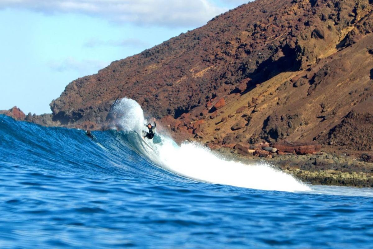 Surfer reitet eine Welle am Ufer mit felsigem, braunem Kliff im Hintergrund.