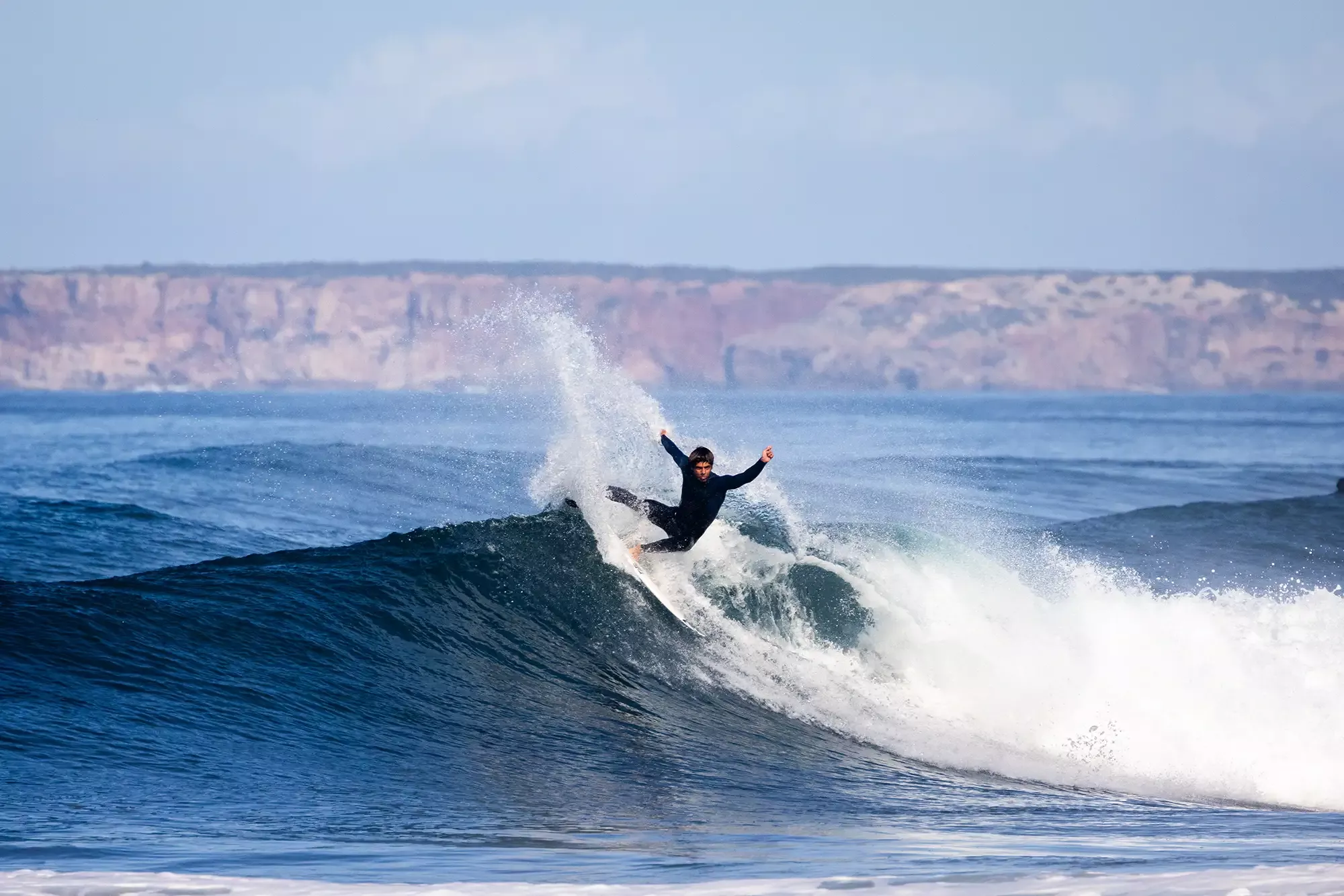 Surfer rides a curling blue-green wave, spraying water as they balance on the board under a clear sky.