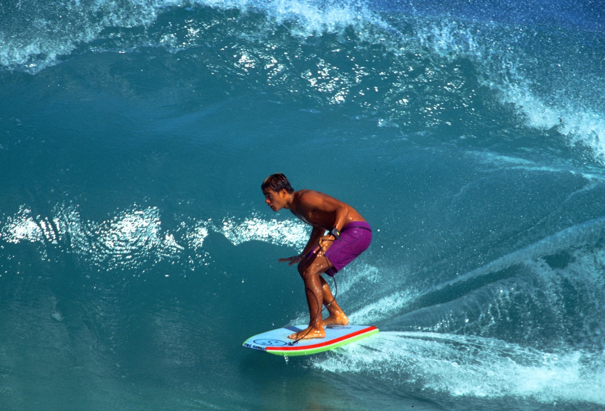 Surfer riding a blue curl, crouched on a colourful board inside a breaking turquoise wave.