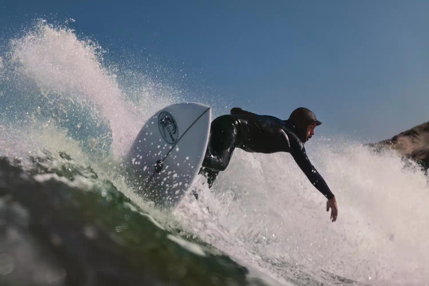 Surfer riding a breaking wave, crouched low on a white shortboard as spray erupts around.