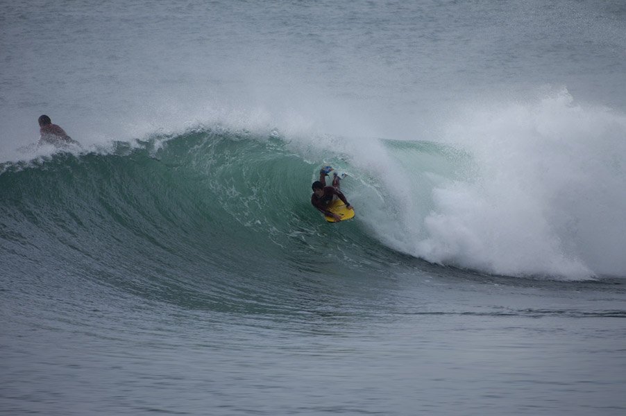 Surfer riding a green barreling wave on a bright yellow board, with spray behind. A second person swims in the background.