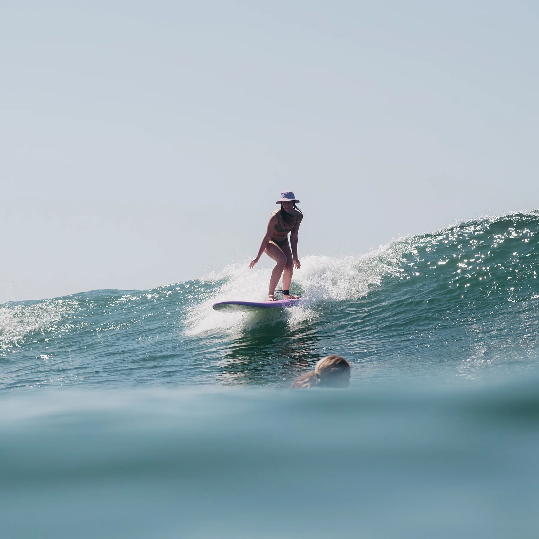Surfer riding a small wave on a longboard, wearing a hat and swimsuit, with another person’s head just underwater in the foreground.