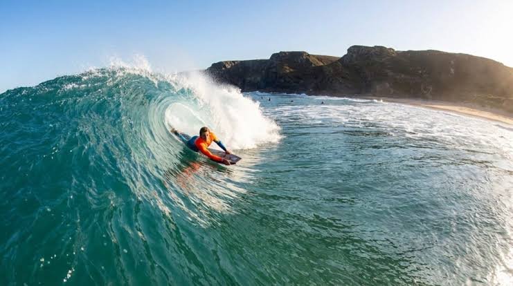 Surfer riding a turquoise wave inside a barrel near rocky coastline, sunny sky.
