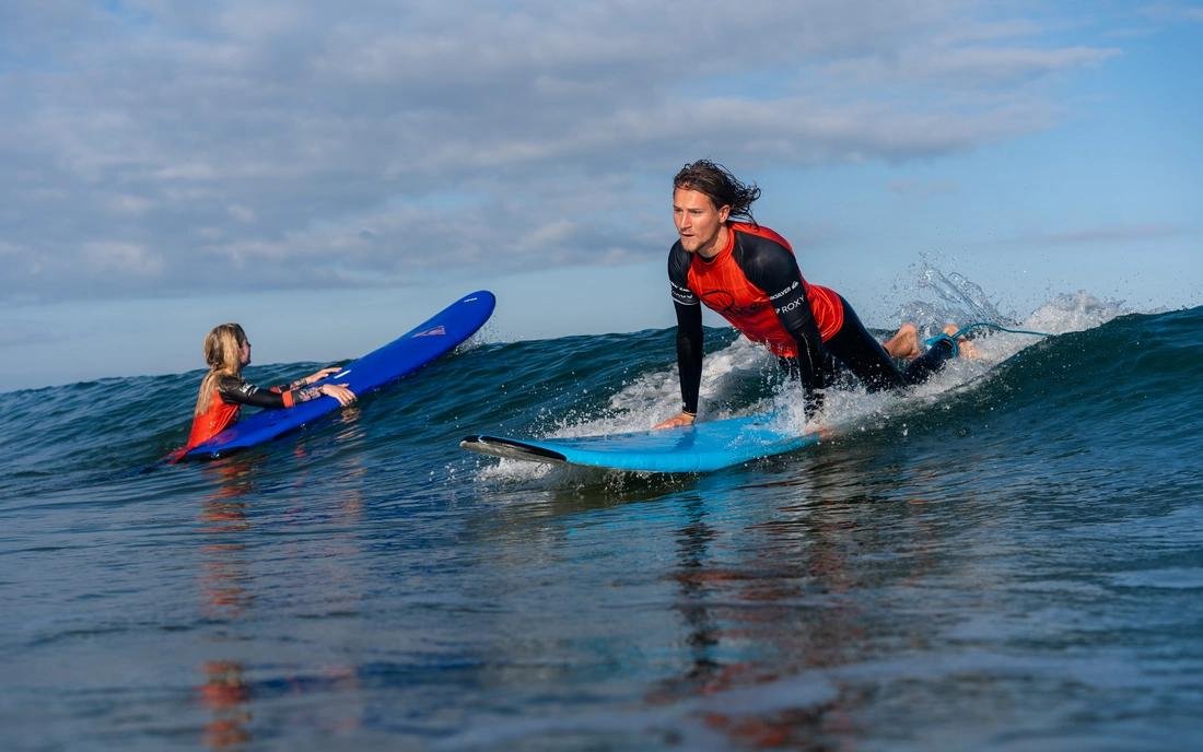 Surfer in rotem Neoprenanzug paddelt auf einer Welle, ein weiterer Surfer liegt auf dem Surfbrett dahinter; klares Blau des Meeres und Himmel.
