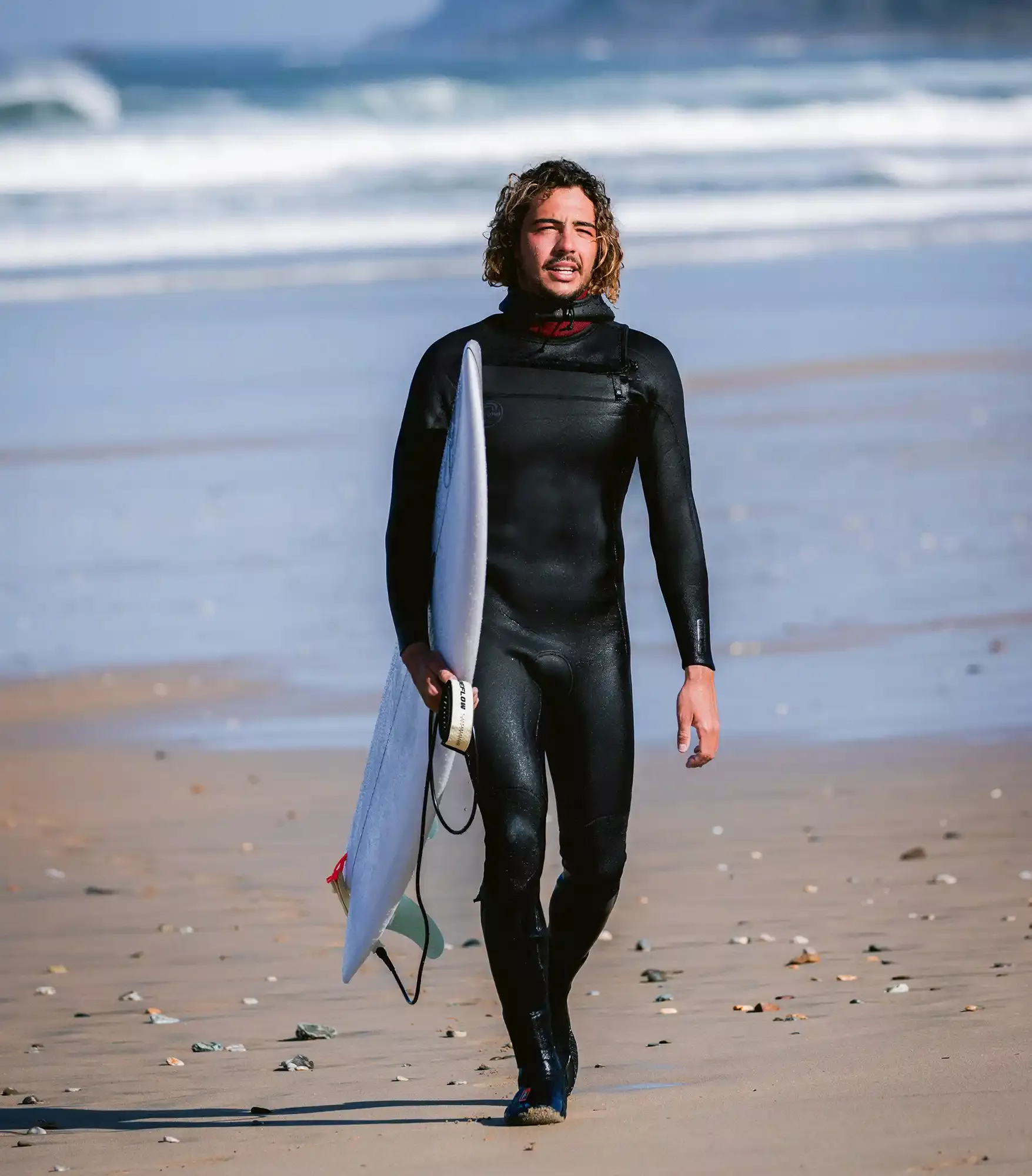 Surfer in schwarzem Neoprenanzug mit Surfbrett unter Arm läuft am Strand entlang, Meereswellen im Hintergrund.