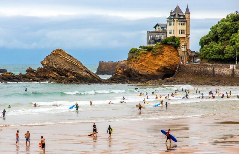 Surfer am Strand vor felsiger Küste und einem sakralen Turmgebäude auf Klippen, gemütliche Welle.