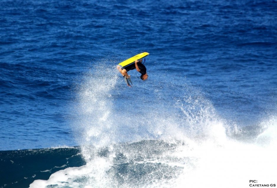 Surfer stürzt mit gelbem Bodyboard in einem Sprung über eine hohe Welle ins Wasser.