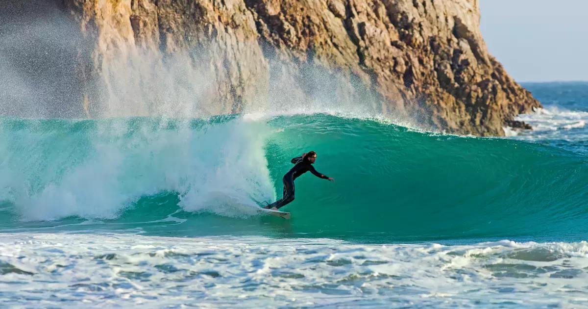 Surfer auf einer türkisfarbenen Welle in einer Küstenbrandung, vor sich eine hohe Wasserwand.