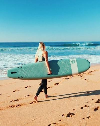 Surfer walking along sandy beach with a mint-green surfboard under arm, Ocean waves in background.