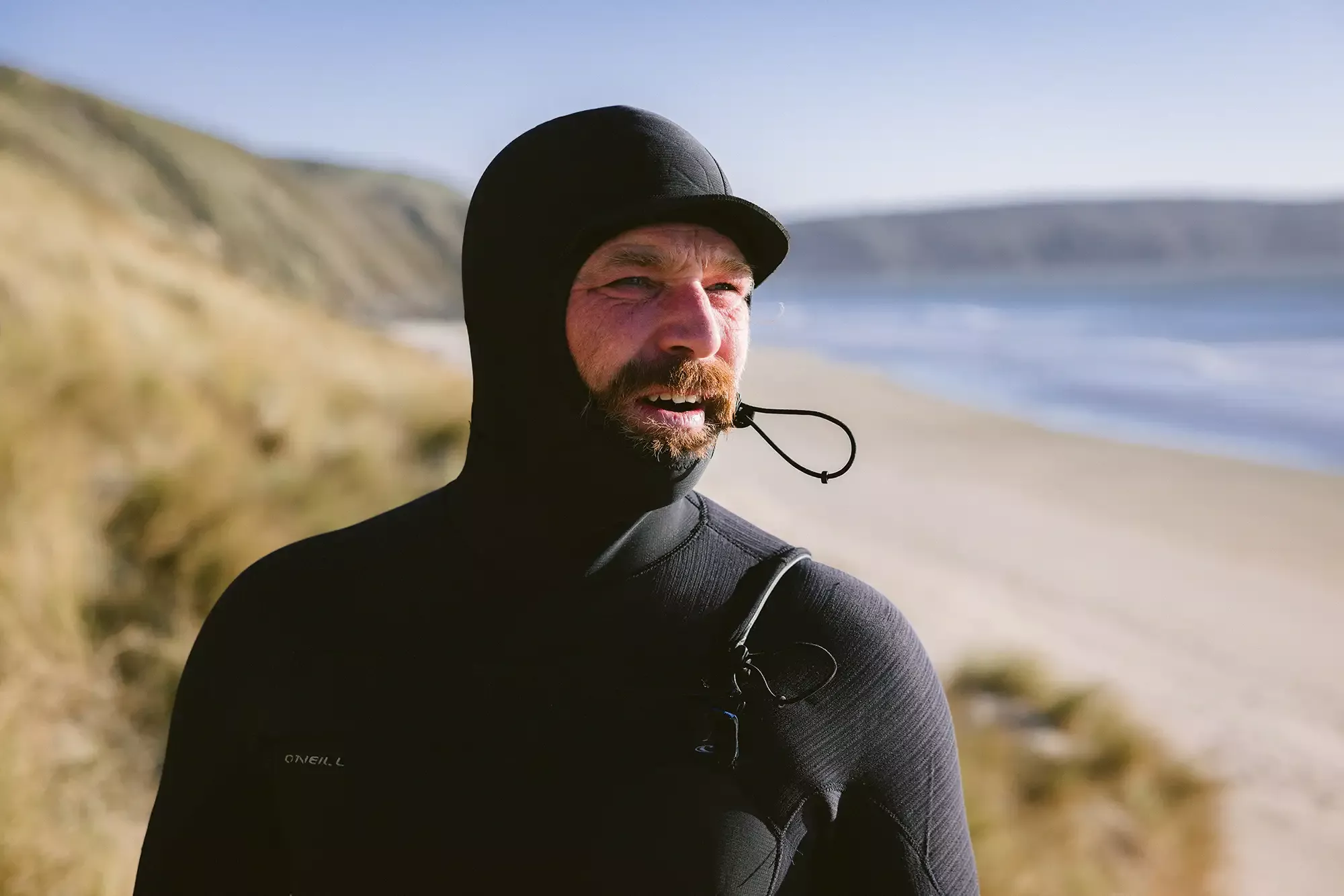 Surfer wearing a black wetsuit hood with a strap, standing on a sandy beach with cliffs and blue sea in the background.