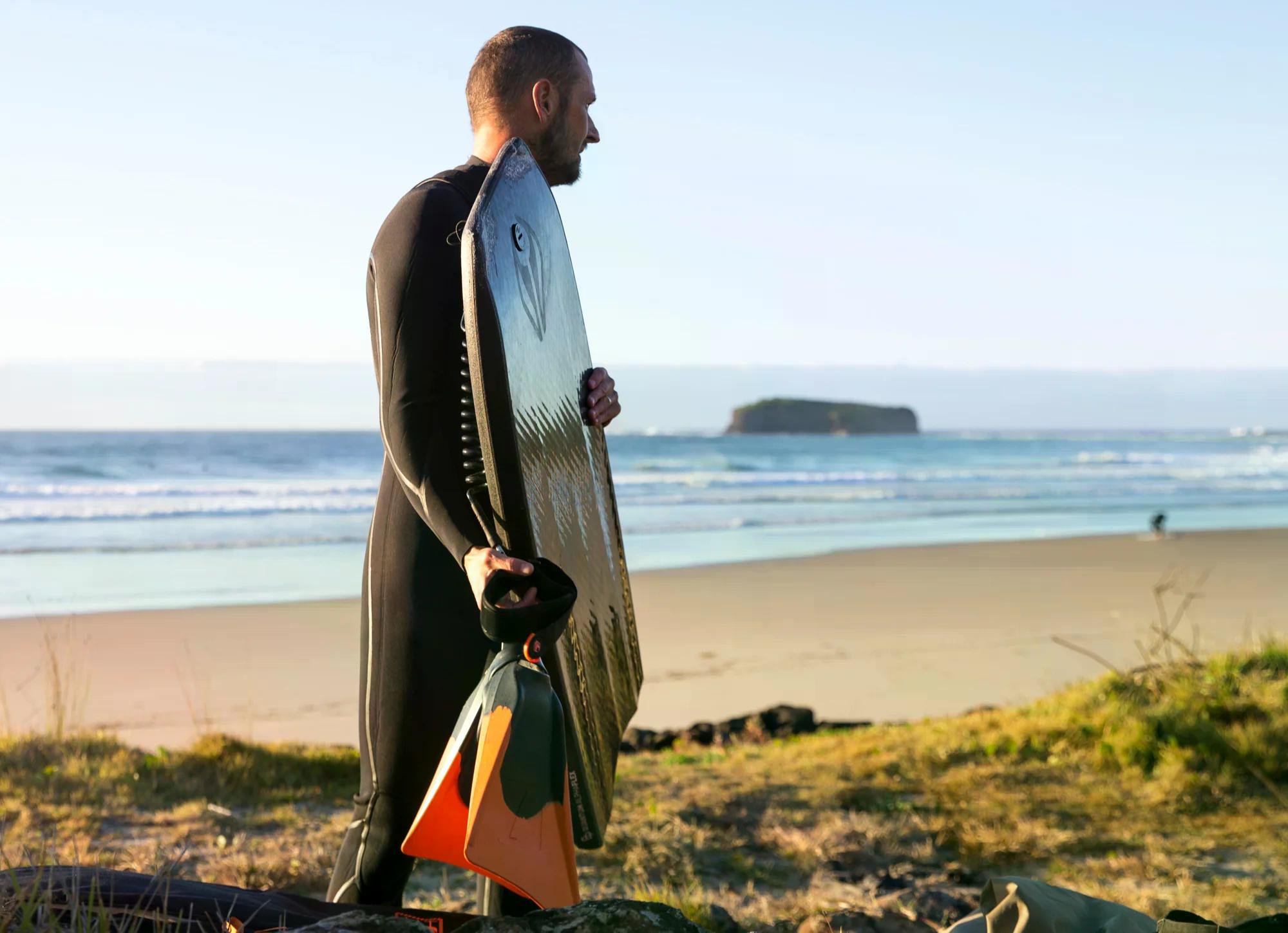 Surfer in wetsuit carrying a large surfboard and a small finned board on a sandy beach.
