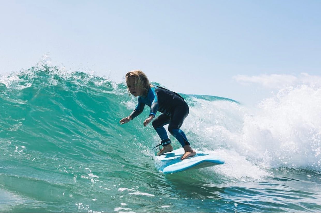 Surferin in Neoprenanzug reitet auf einer Welle mit Surfbrett, während Wasser spritzt und klare blaugrüne See zu sehen ist.