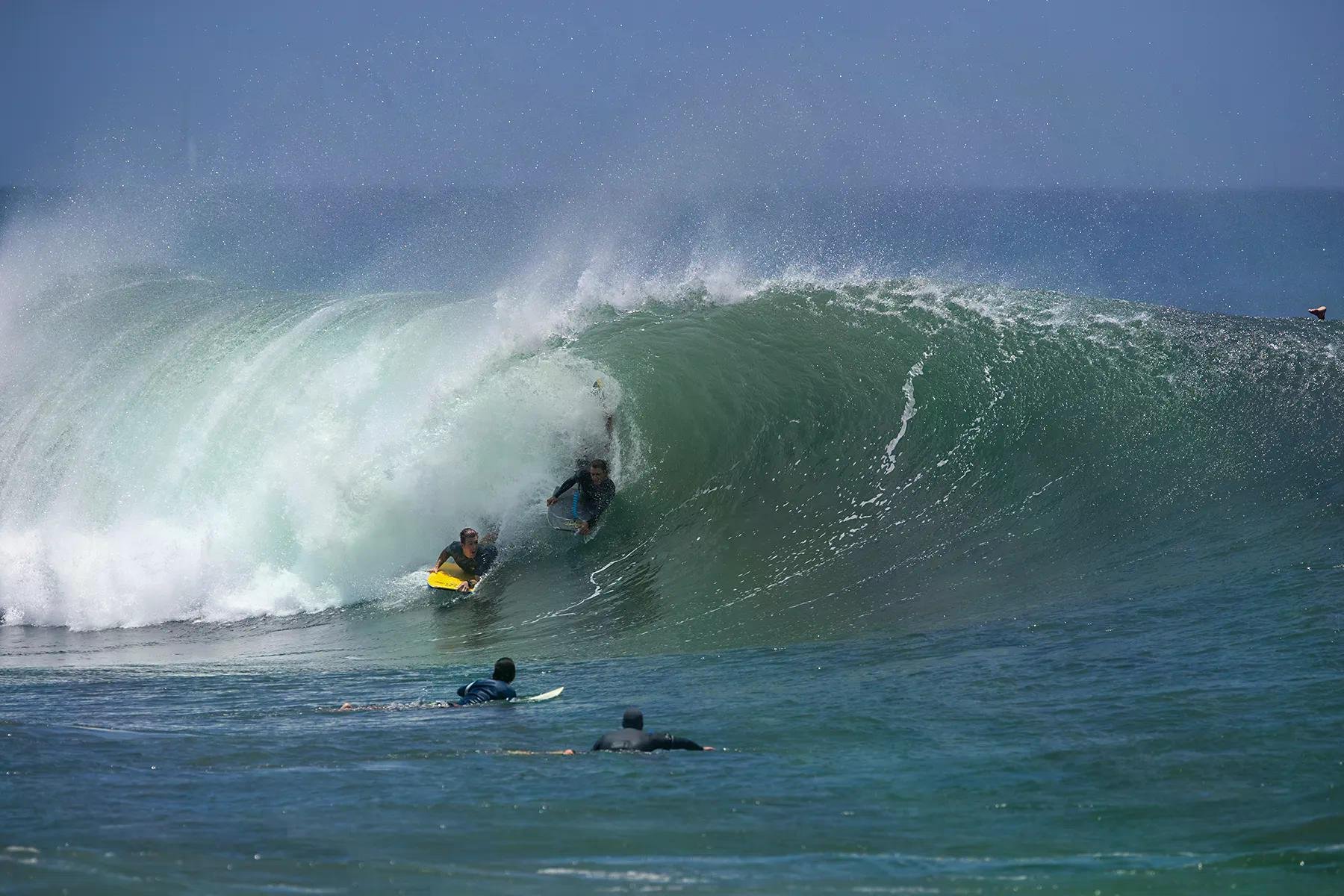 Surfers ride a large green wave, two inside the barrel while others paddle nearby in the foreground.