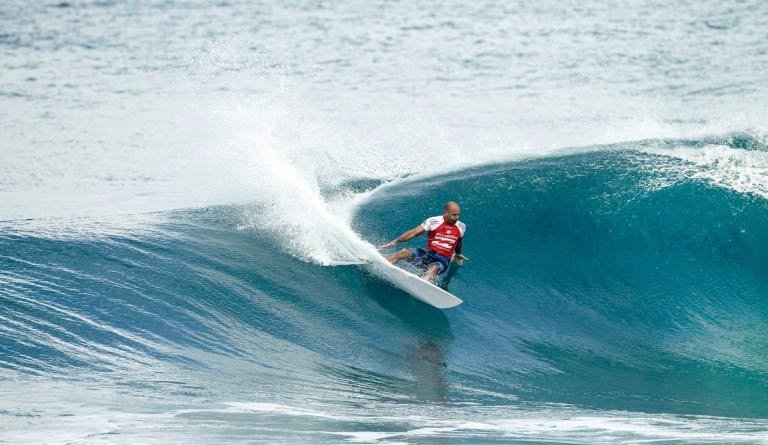 Surfeur à pic de vague, portant gilet rouge, sur une planche dans une grande houle bleue.