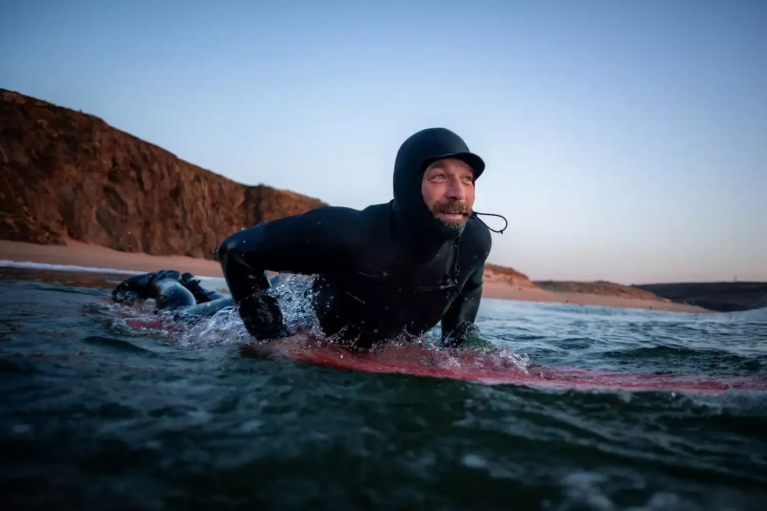 Surfeur en combinaison néoprène noir sur une planche, nageant debout dans l’eau près d’une plage rocheuse au coucher du soleil.