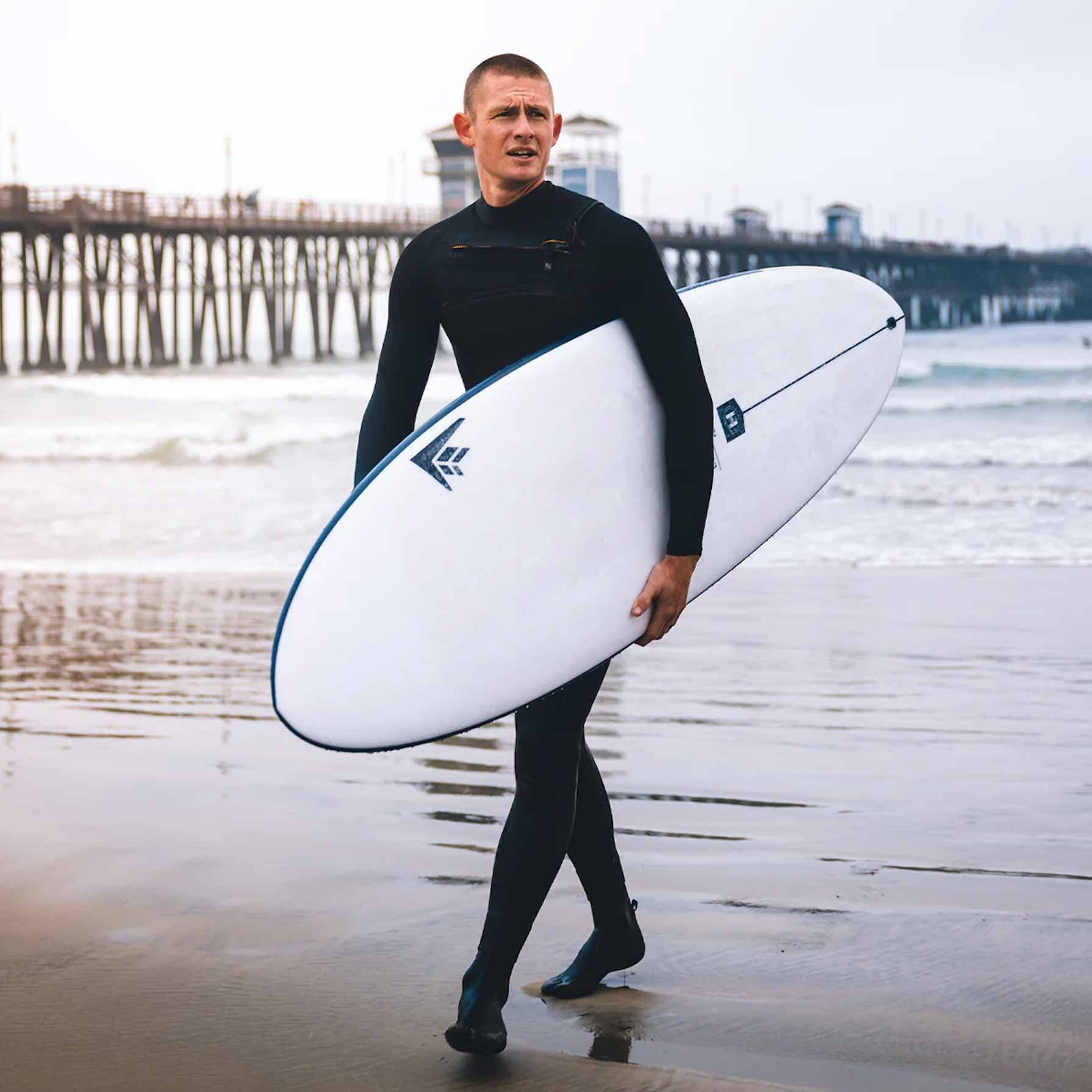 Surfeur en combinaison noire marche sur la plage avec une planche de surf blanche sous le bras, près de l’eau et d’un quai en arrière-plan.