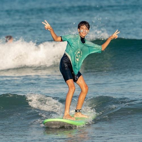 Surfeur jeune sur une planche de surf verte, bras levés, ride sur une petite vague au bord de l’océan.