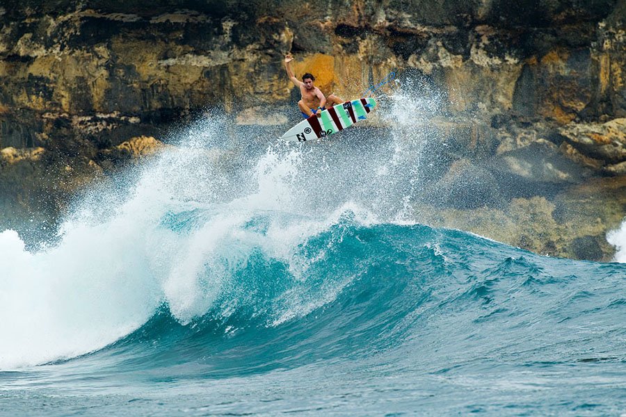 Surfeur sur une planche, en vol au-dessus d’une vague turquoise près de falaises rocheuses.