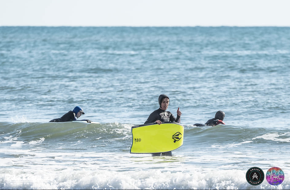 Surfeur tenant une planche jaune fluorescent dans une petite vague, en mer avec deux autres surfeurs à l’arrière; logos et badges de partenaires en coin.