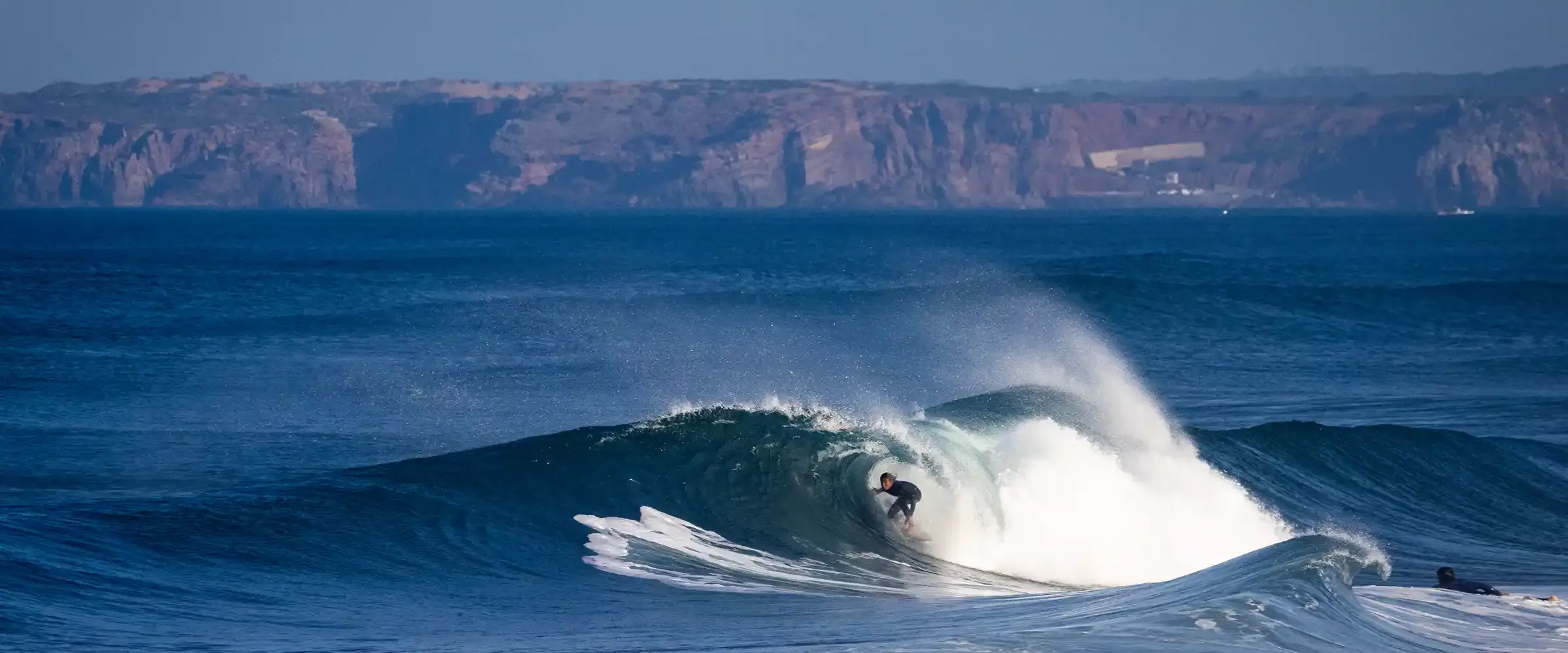 Surfista cabalgando una ola en mar abierto, con cresta blanca y fondo de acantilados.