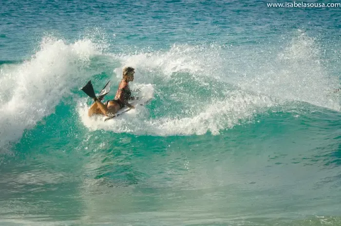 Surfista a cair numa onda azul, com splash de água perto da arrebentação.