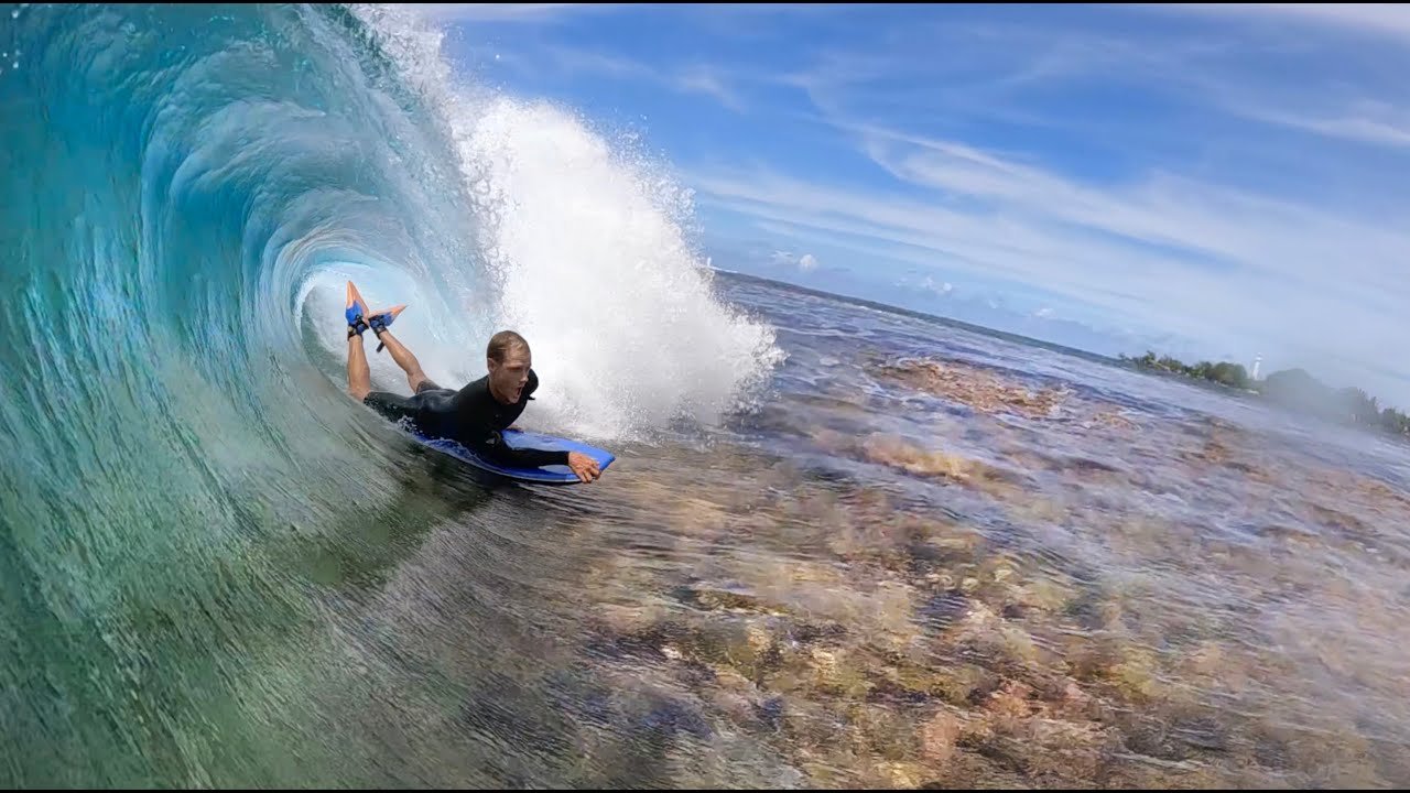 Surfista dentro de una ola azul, rompiendo en una pared amplia, con el cuerpo apoyado sobre una tabla y agua clara alrededor.