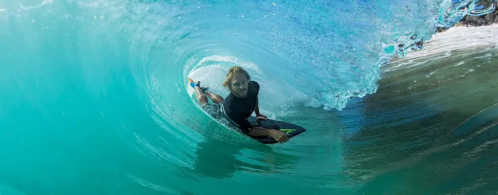 Surfista dentro de una ola hueca, rodeado de agua azul translucido, sosteniendo una tabla de surf mientras avanza en el tubo.