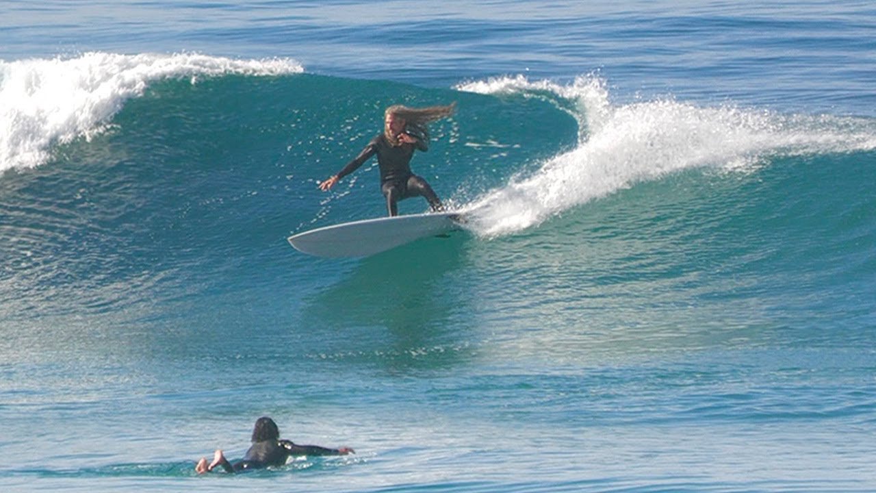 Surfista in equilibrio sulla tavola mentre cavalca un’onda, con un altro nuotatore sullo sfondo.