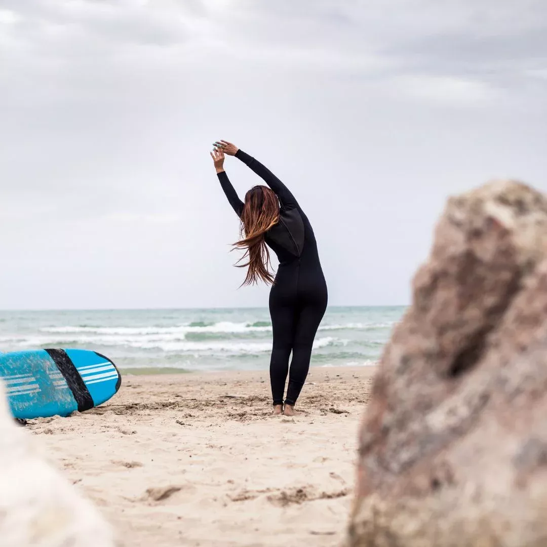 Surfista de espalda en playa, con traje neutro, estirándose junto a una tabla de surf azul en el suelo.