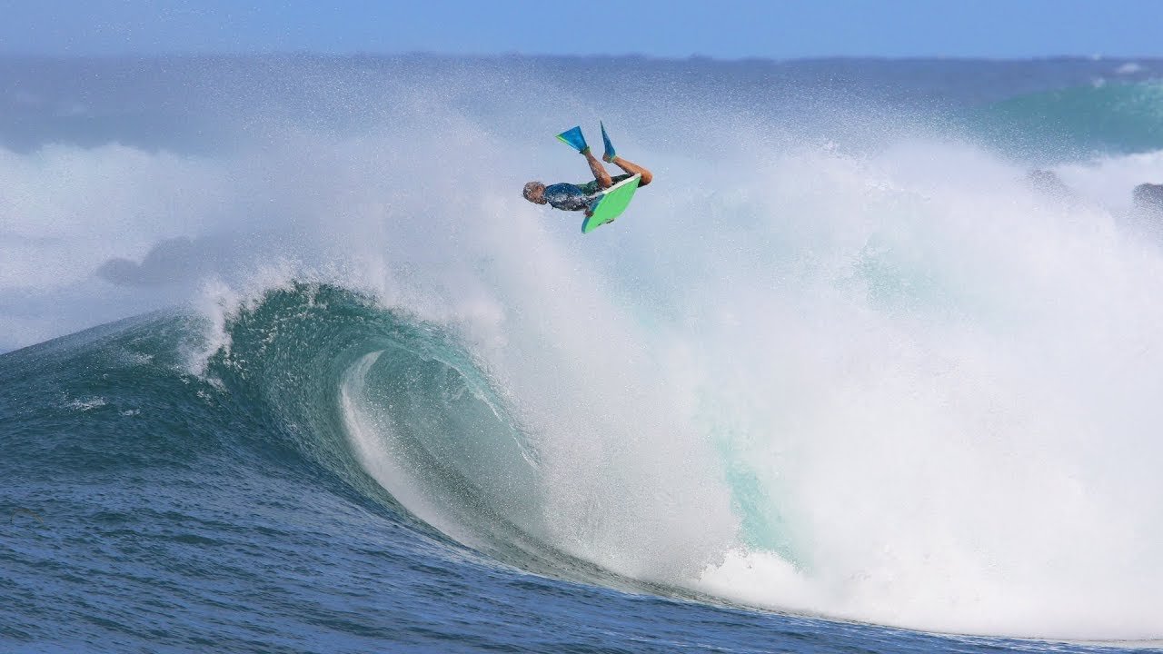 Surfista é lançado ao ar ao pegar uma onda gigante, com a prancha verde vibrante e a água espumando ao redor.
