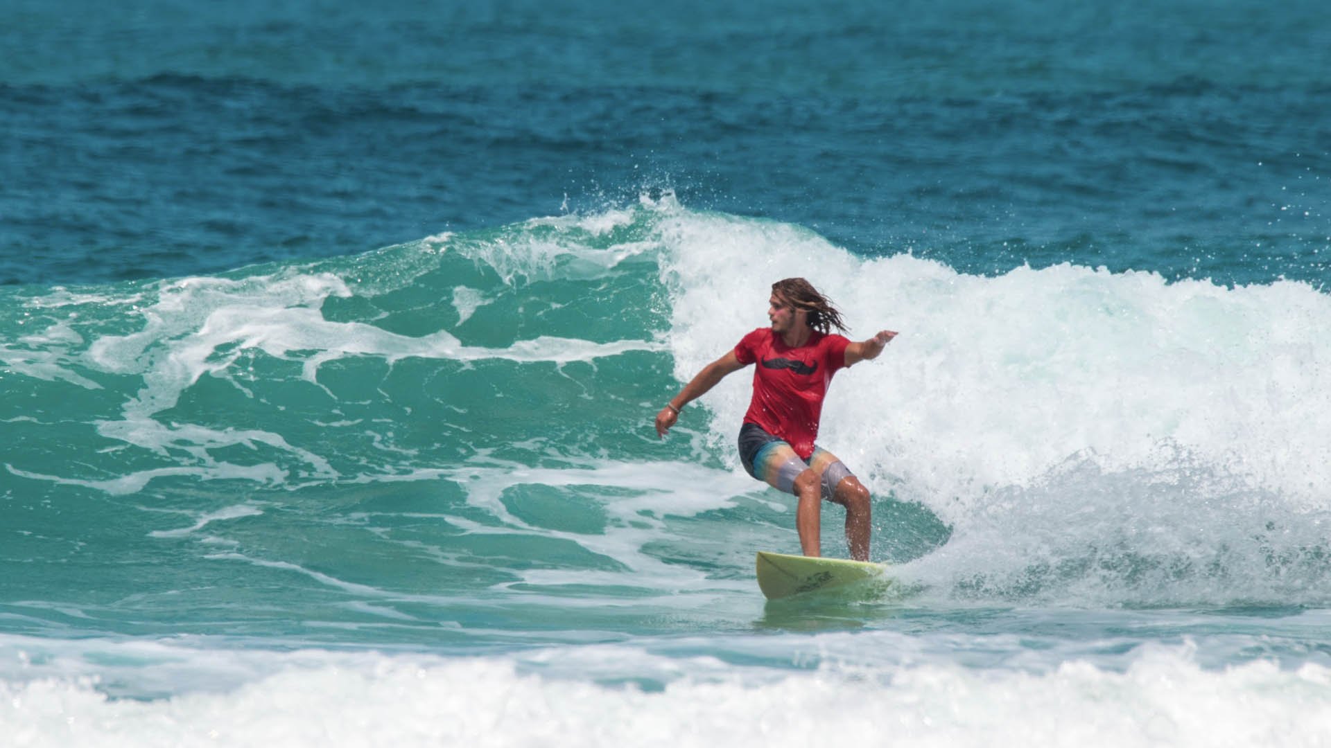Surfista in maglietta rossa su tavola, cavalca una cresta di onda verde-azzurra in mare.