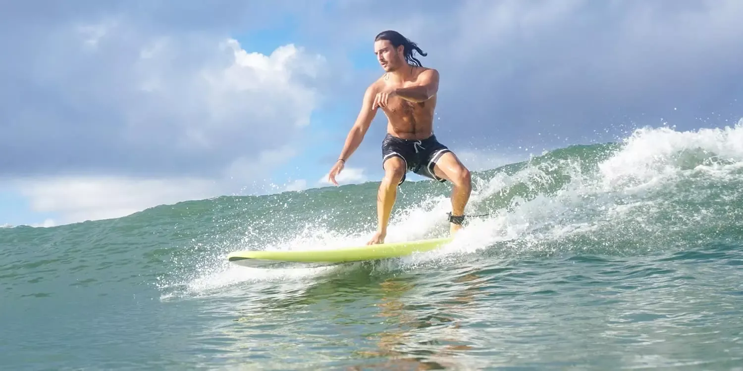 Surfista masculino de pie sobre una tabla de surf en una ola, con el mar y cielo nublado de fondo.