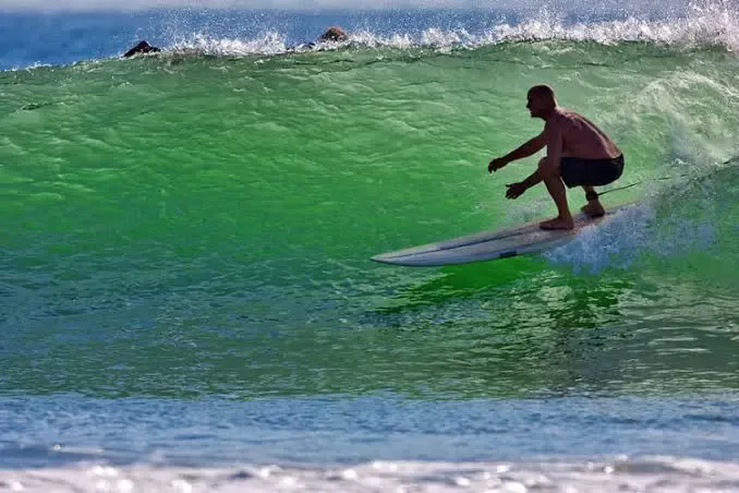Surfista montando una ola verde en mar abierto, deslizándose sobre una tabla corta en posición crouched.