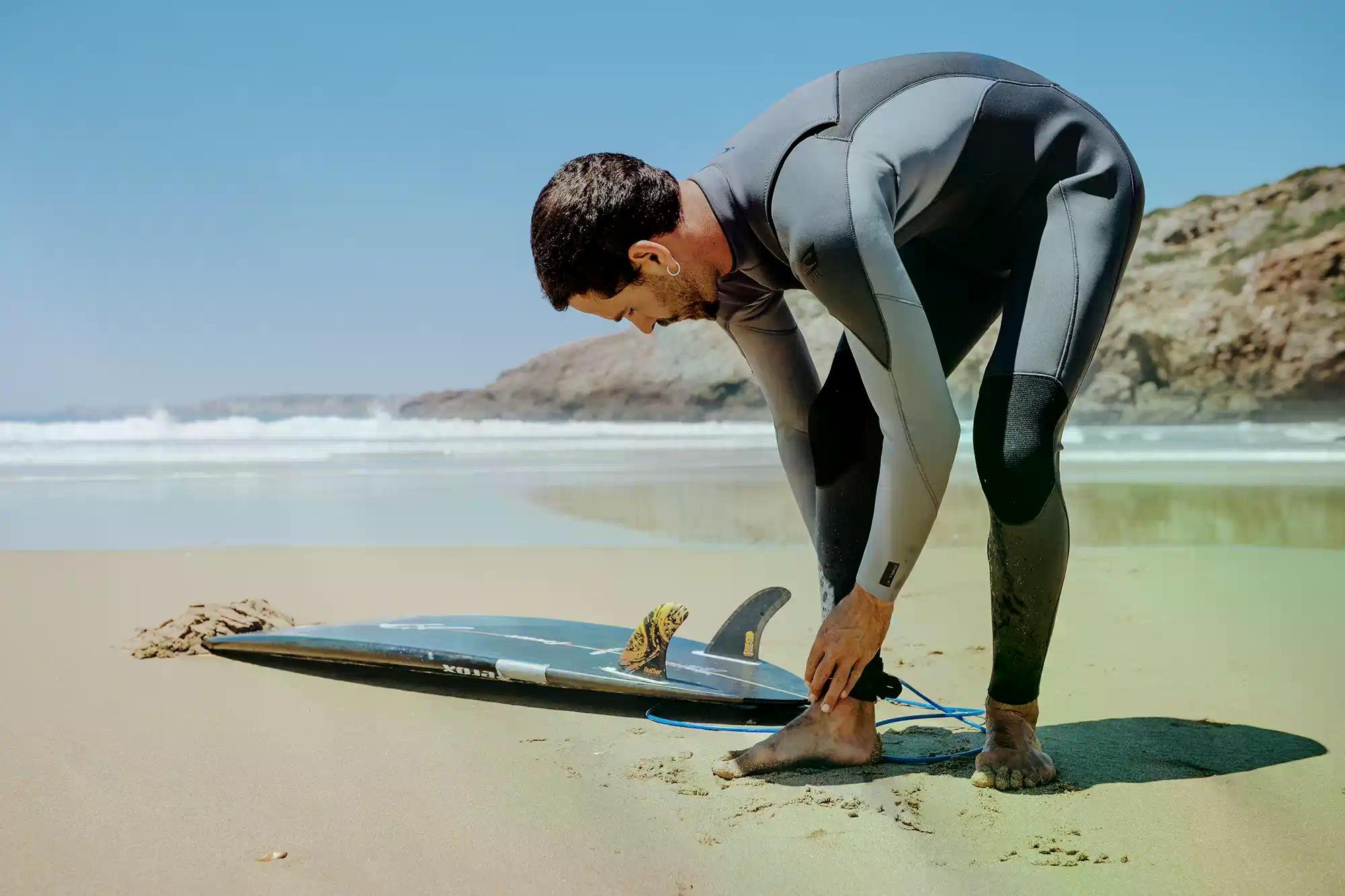 Surfista in muta piegato sul bagnasciuga, sistemando la tavola da surf sulla sabbia rocciosa.