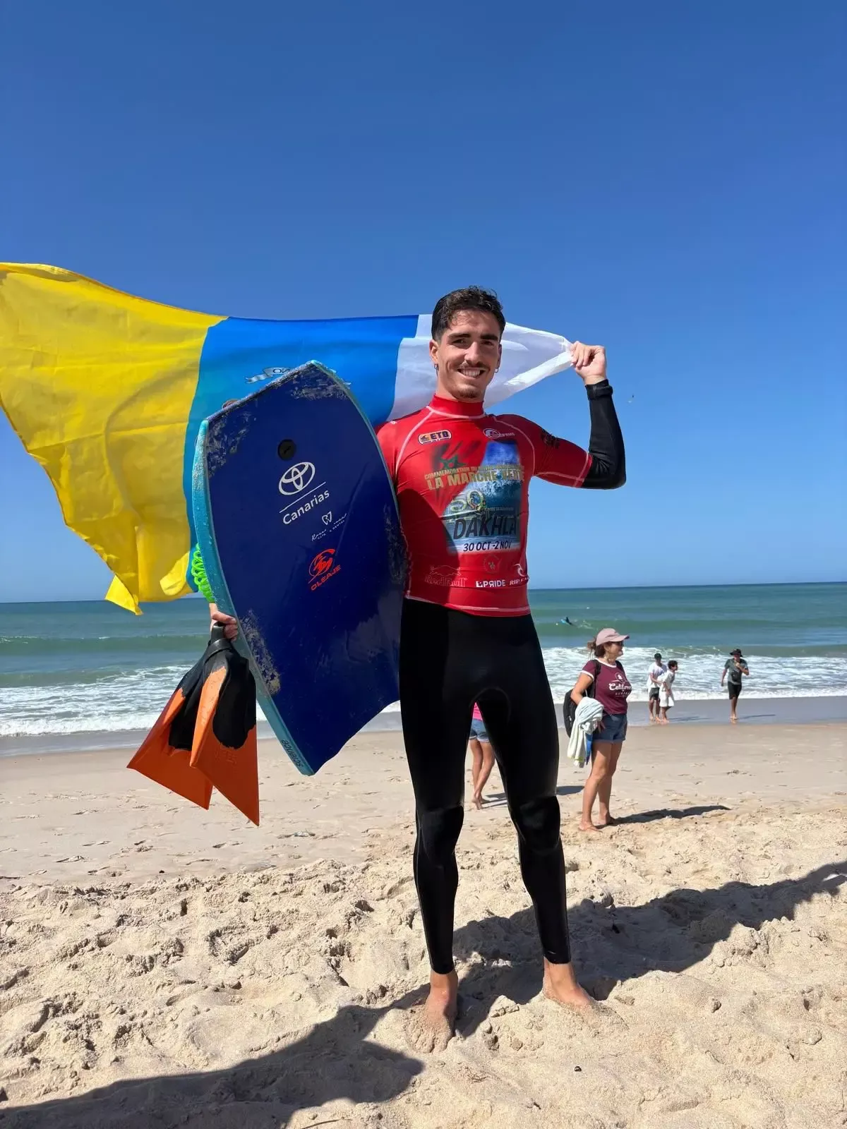 Surfista com prancha azul na prancha, segurando bandeira amarela e azul na praia ensolarada.