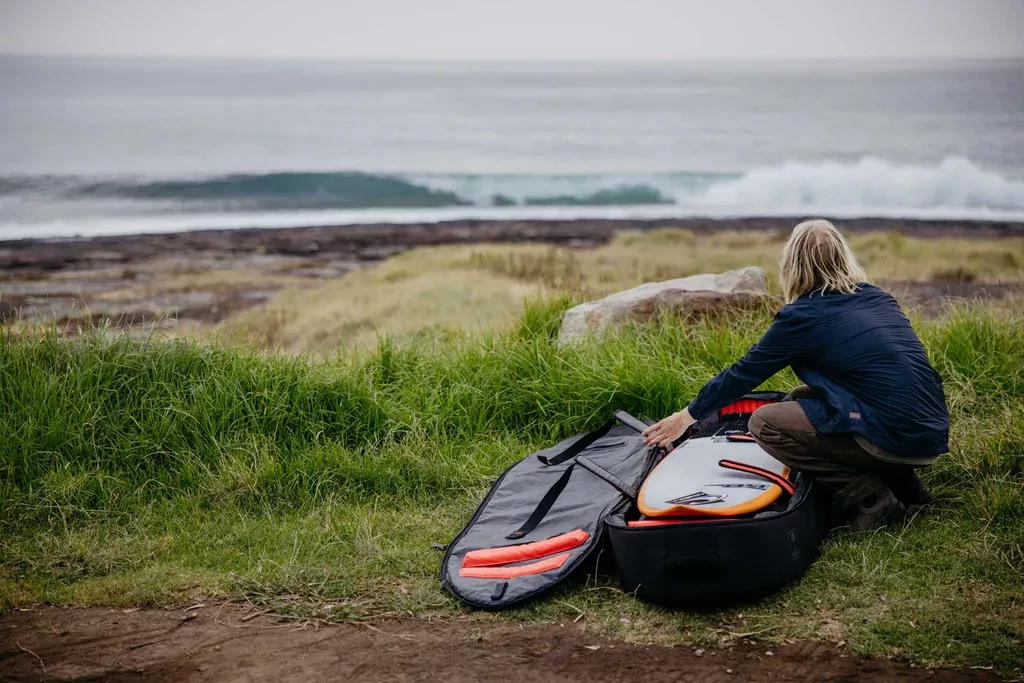 Surfista preparando su tabla de surf en un entorno costero con maleta y equipo, lista para montar gradería y olas.