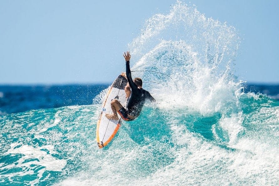 Surfista realizando maniobra sobre una ola en una tabla, con salpicaduras de espuma y mar azul.