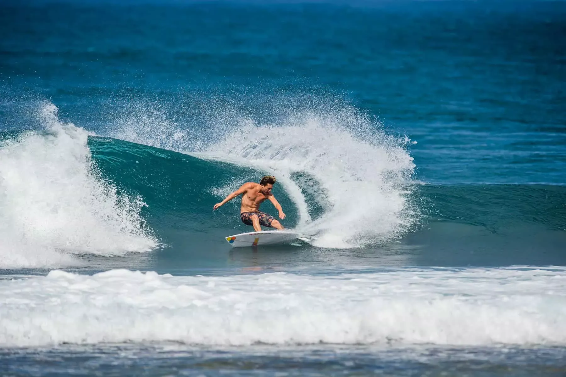 Surfista en tabla corta dentro de una ola curva, spray blanco y mar azul intenso de fondo.