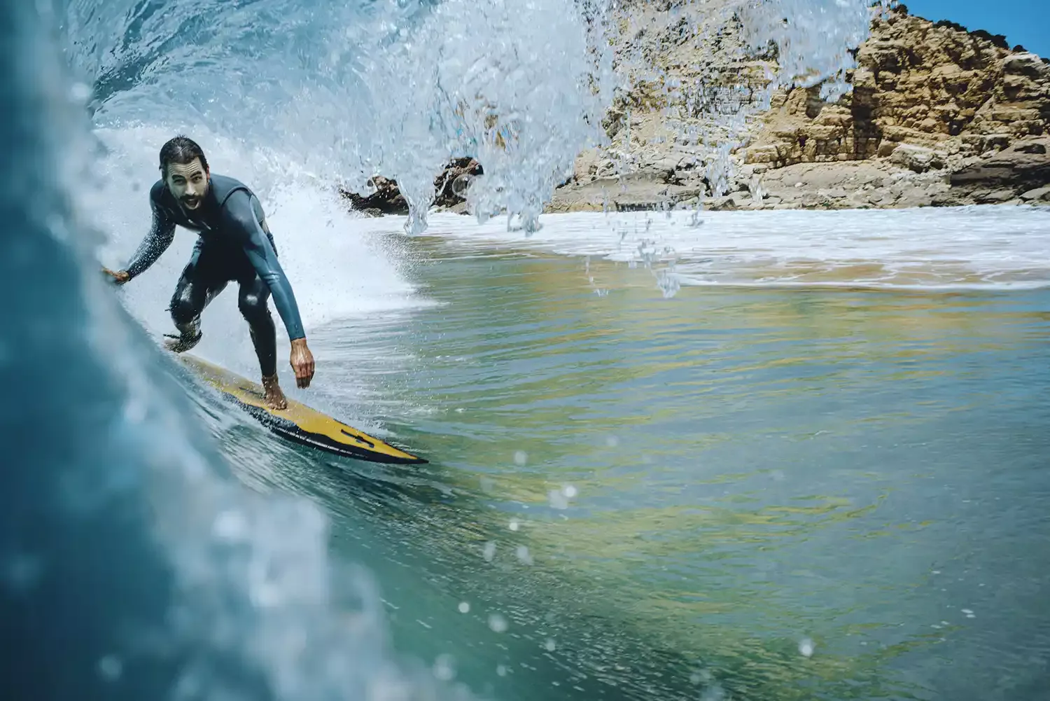 Surfista sobre una tabla en una ola rodeada de agua clara y formaciones rocosas al fondo.