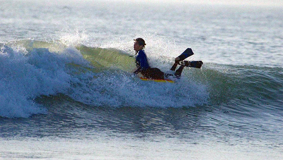 Surfista su tavola corta in mezzo alle onde, con pinne e torso neutro, pronto a rialzarsi. Altamente dinamico.