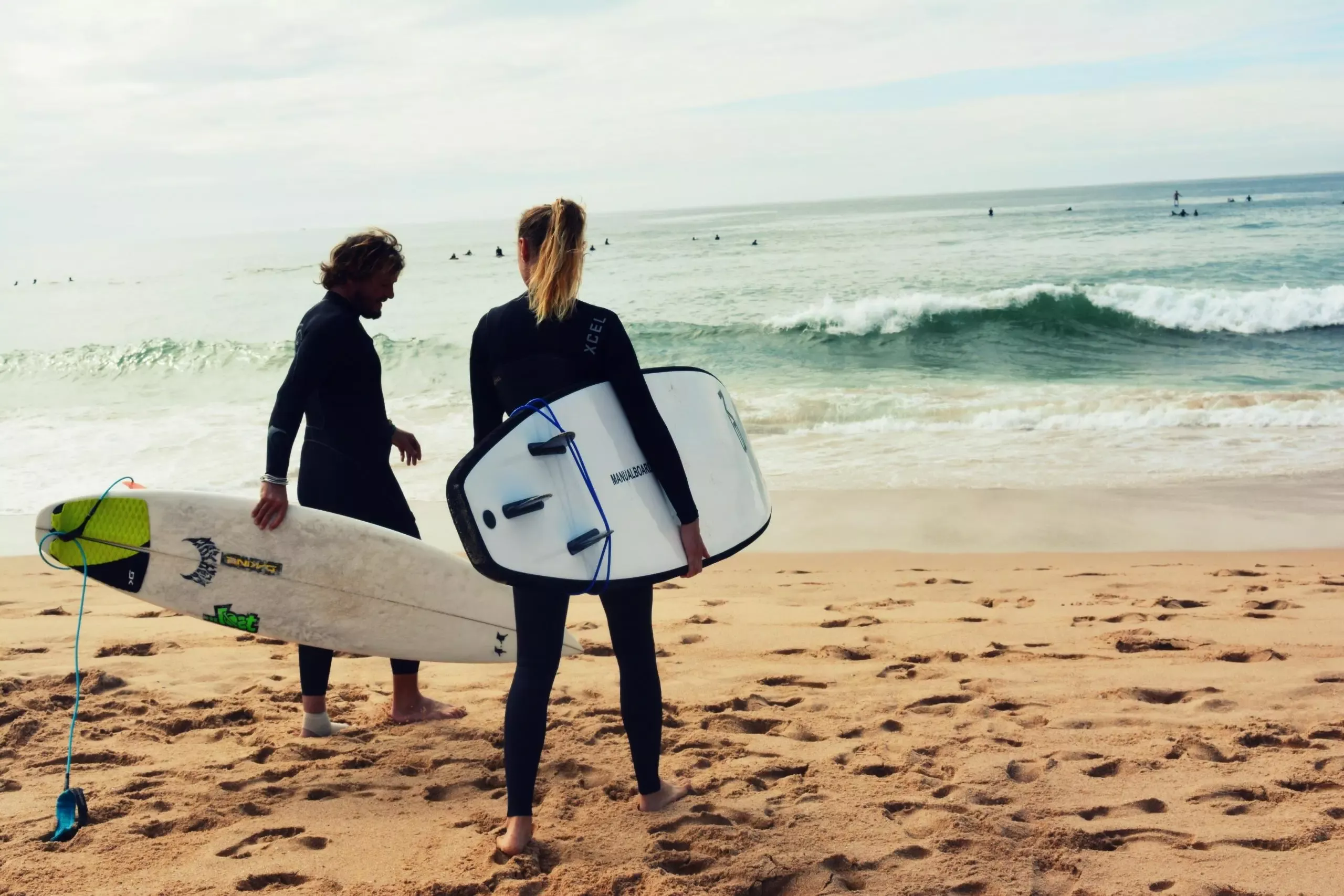 Surfistas à beira-mar com prancha de surf na areia, prontos para entrar no oceano.