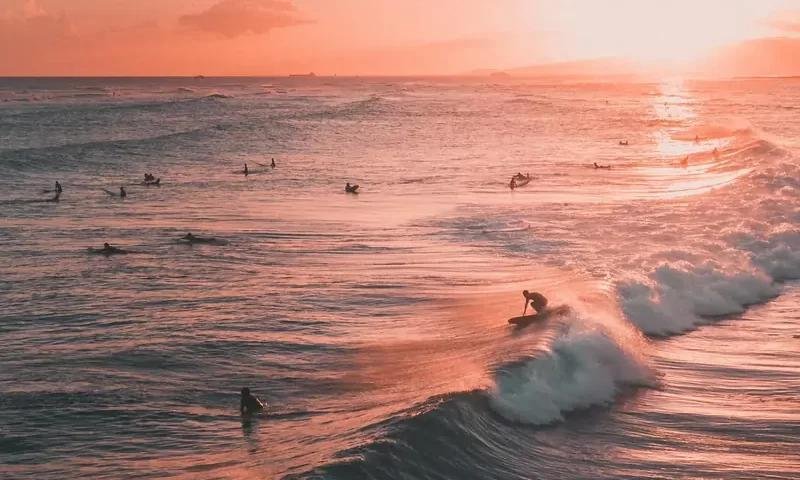 Surfisti in mare al tramonto, onde rosa e azzurre si infrangono, scena di beach session.
