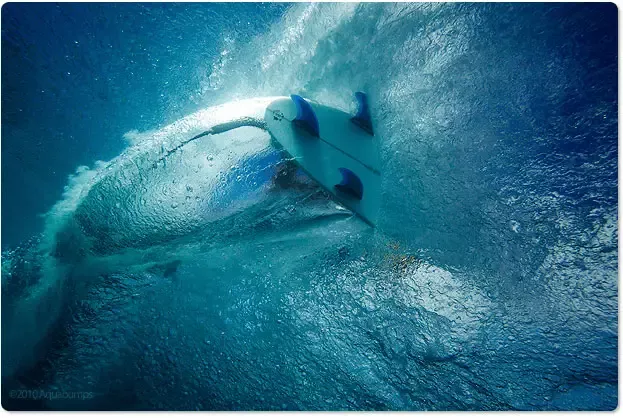 Tabla de surf curva dentro de una ola azul, rodeada de espuma y agua cristalina.