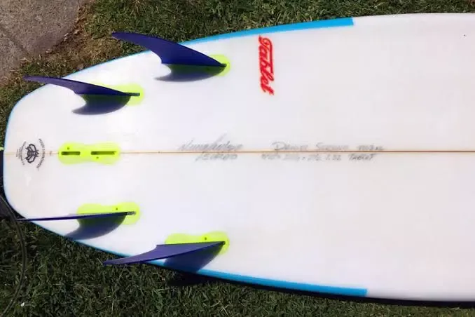 Tablero de surf visto de arriba con aletas azules y amarillas, y quillas en punta; diseño blanco con acentos azules y detalles rojos.