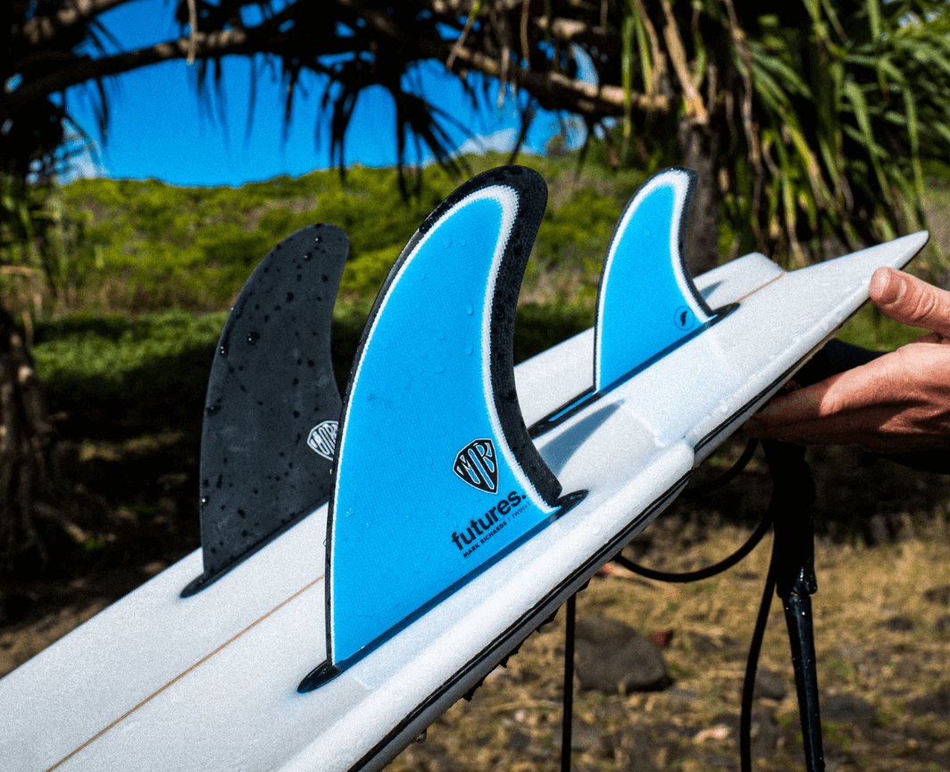 Tres aletas de surf azul y negra apoyadas en una tabla, con una mano sujetando la cubierta; fondo natural con césped y árboles.