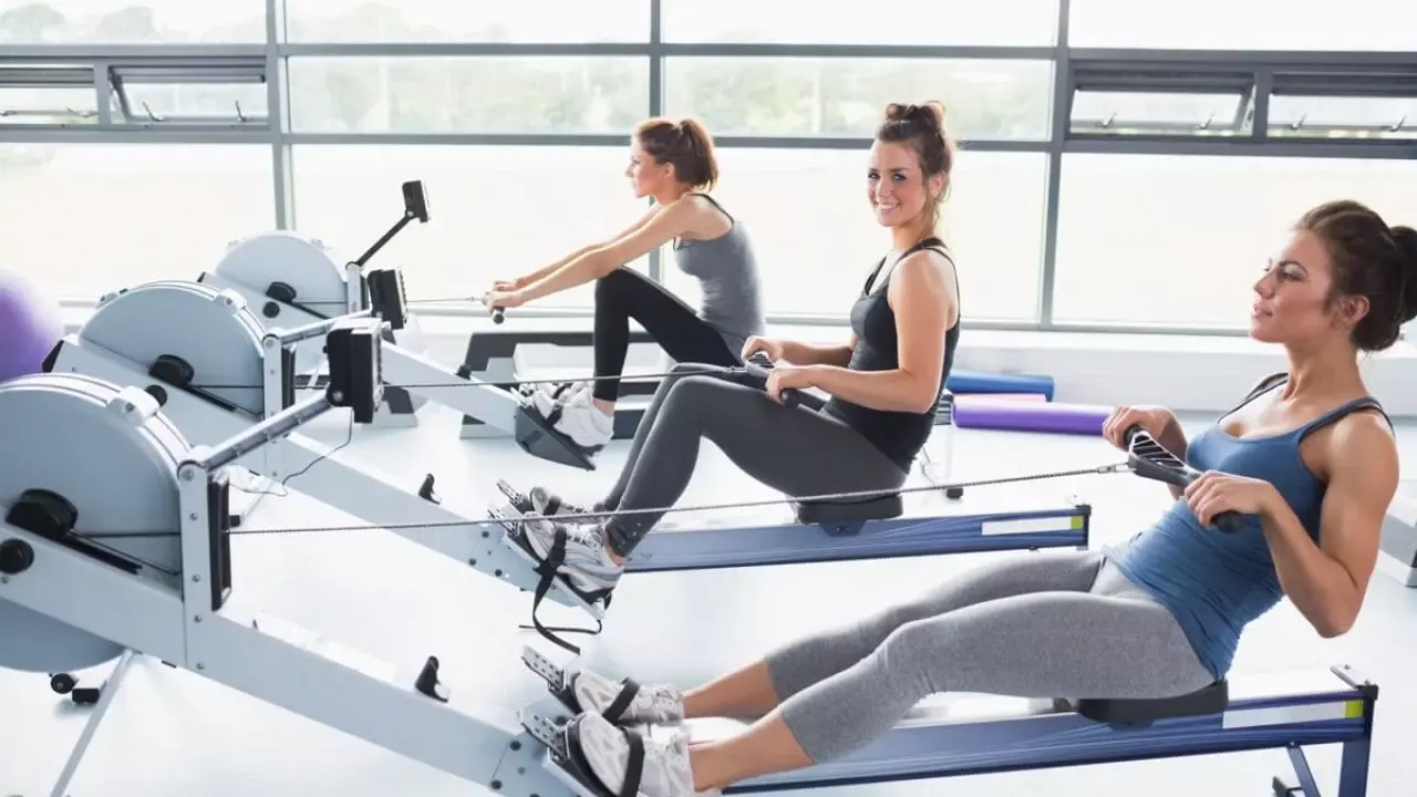 Tres mujeres ejercitando en una sala con máquinas de remo, sonriendo y mirando al frente. Están con ropa deportiva y pantorrillas recostadas en las máquinas.