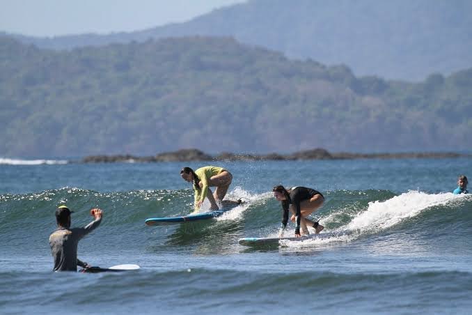 Três surfistas em prancha, pegando onda na água com montanhas ao fundo.